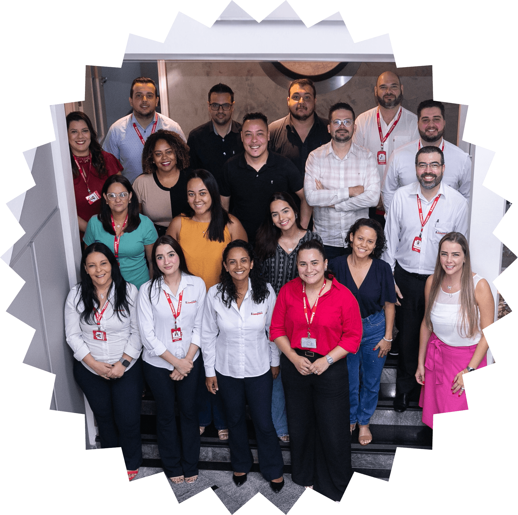 Um grupo de dezoito pessoas está reunido para uma foto em um ambiente interno, perto de uma escada. A maioria está de pé, enquanto algumas pessoas estão sentadas nos degraus. Todos estão sorrindo para a câmera. Várias pessoas usam crachás vermelhos, e algumas vestem camisas brancas com o logotipo da 'Junqueira'. Há uma mistura de homens e mulheres, e o ambiente é iluminado, com uma área de plantas visível à direita. A atmosfera parece ser de camaradagem e trabalho em equipe.